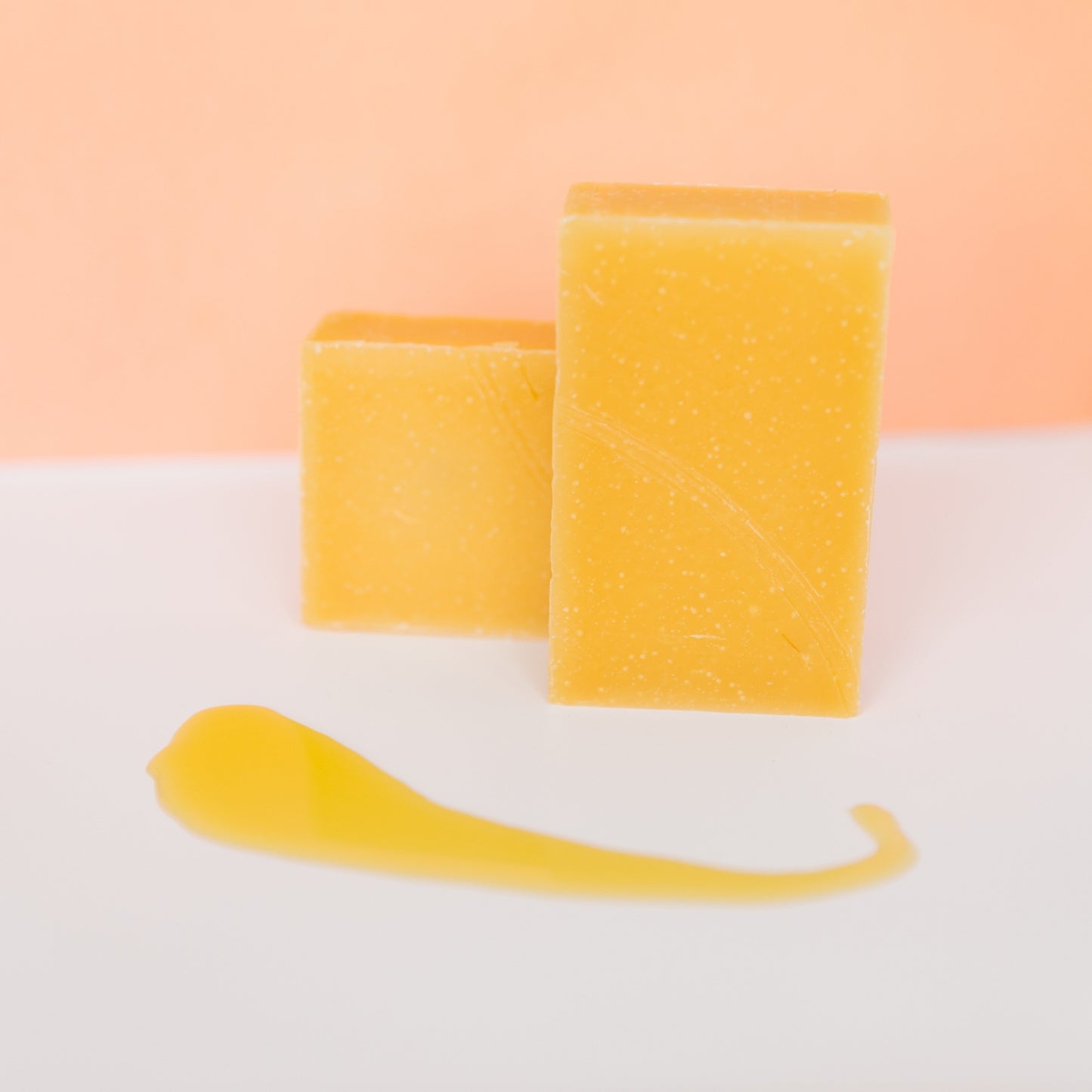Two yellow soaps and a yellow tool on a white surface with a peach-colored background