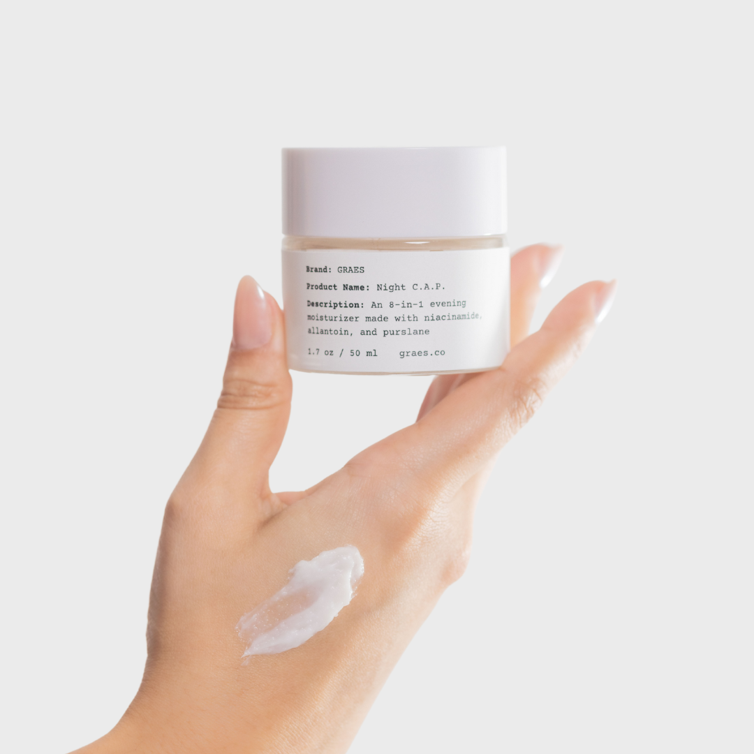 Hand holding a jar of cream with visible cream on the back of hand against a light background
